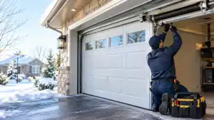 A technician in uniform kneeling by a toolbox while repairing a residential garage door during winter.